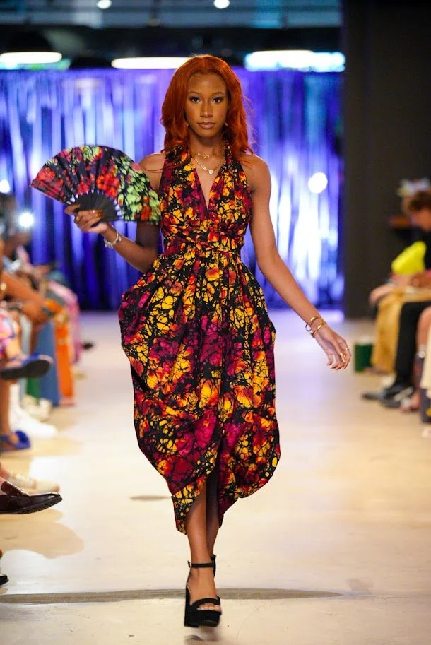 A female model walking down a runway wearing a colorful, patterned dress and holding a hand fan, with an audience on either side of the runway.