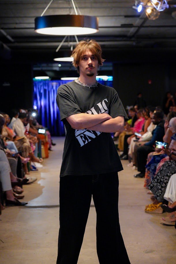Young man with light brown hair and a mustache standing with arms crossed on a runway during a fashion show, surrounded by seated audience members watching and taking photos.