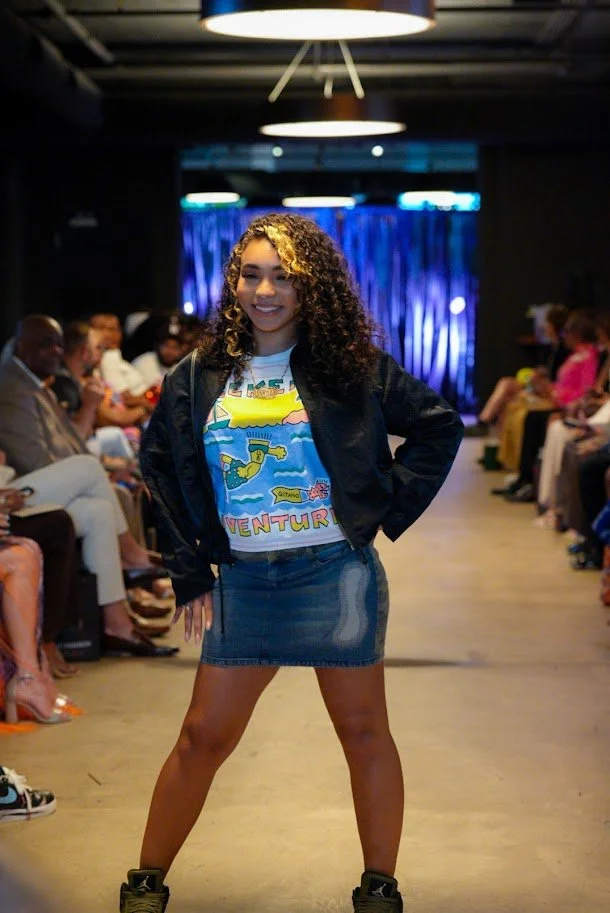 Young woman with curly hair smiling and posing confidently on a runway, wearing a graphic T-shirt, denim skirt, black jacket, and sneakers, with an audience seated on either side of the runway and blue curtain backdrop.