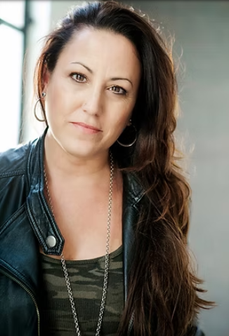 A woman with long brown hair, wearing hoop earrings, a leather jacket, and a striped shirt, looking at the camera.