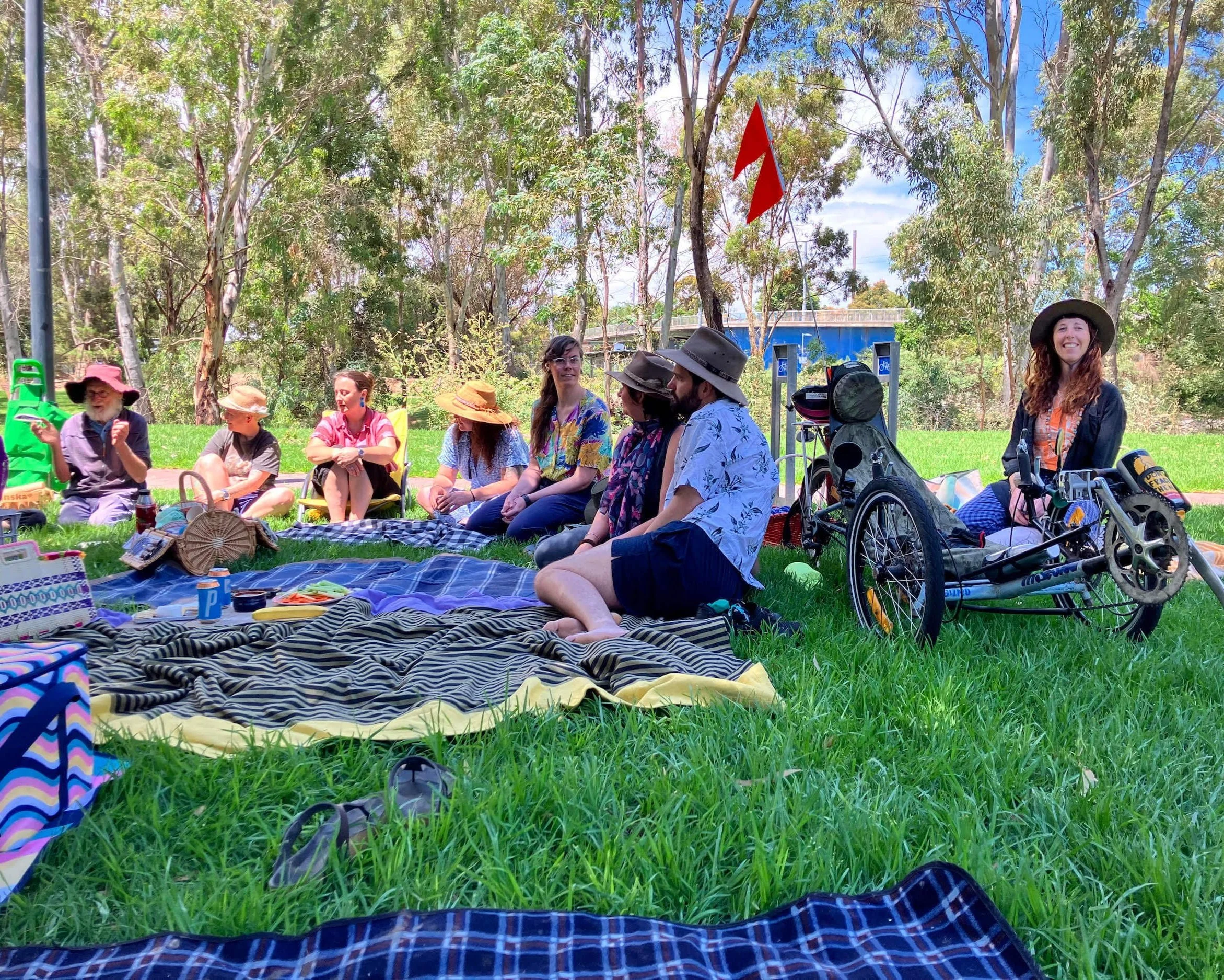 A community picnic with members of all ages sitting on rugs in the shade, enjoying shared conversation and food — become a member and be part of grassroots gatherings like this.