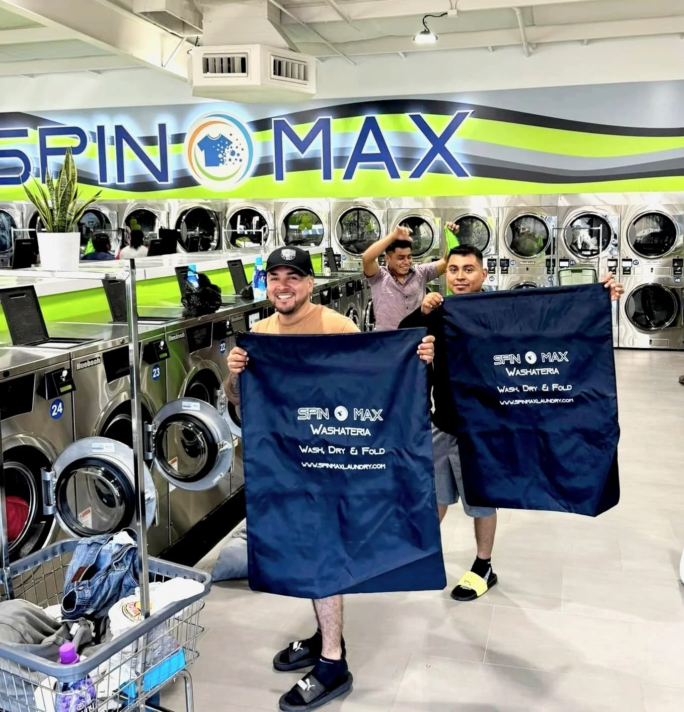 Two people standing in a laundromat holding "Spin Max Washateria" laundry bags. They are smiling, and the background shows washing machines and a sign reading "SPINO MAX". A laundry cart with clothes is in the foreground.