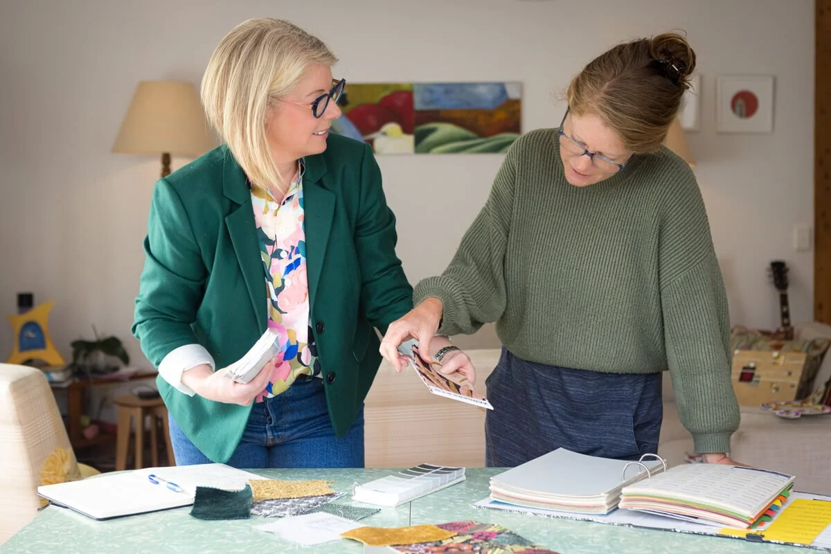 Interior designer Nicci Tong and female client discussing fabric swatches at a table with notebooks and open binders, engaging in a design or planning activity.