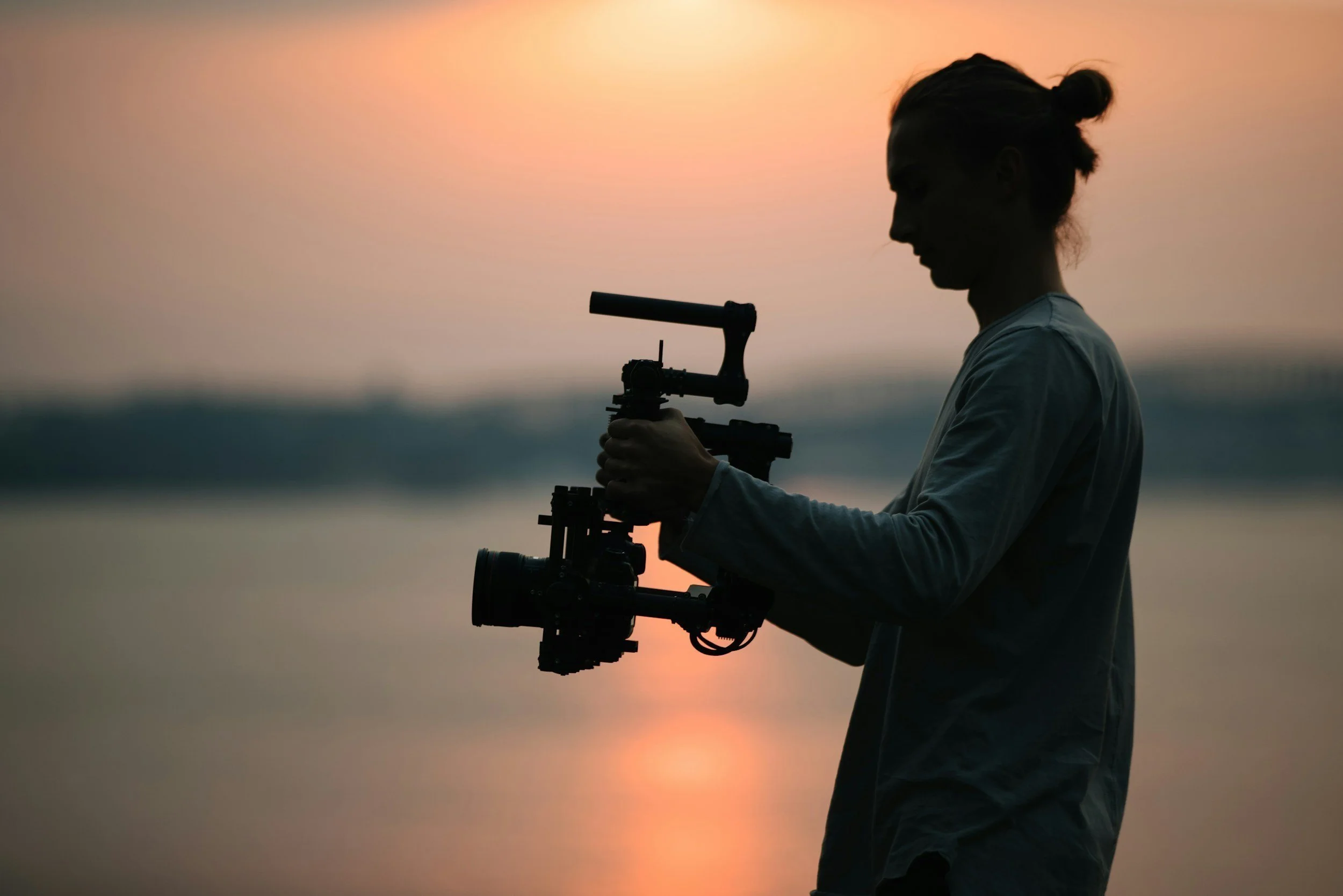 Silhouette of a person holding a professional camera or camera stabilizer during sunset by the water.