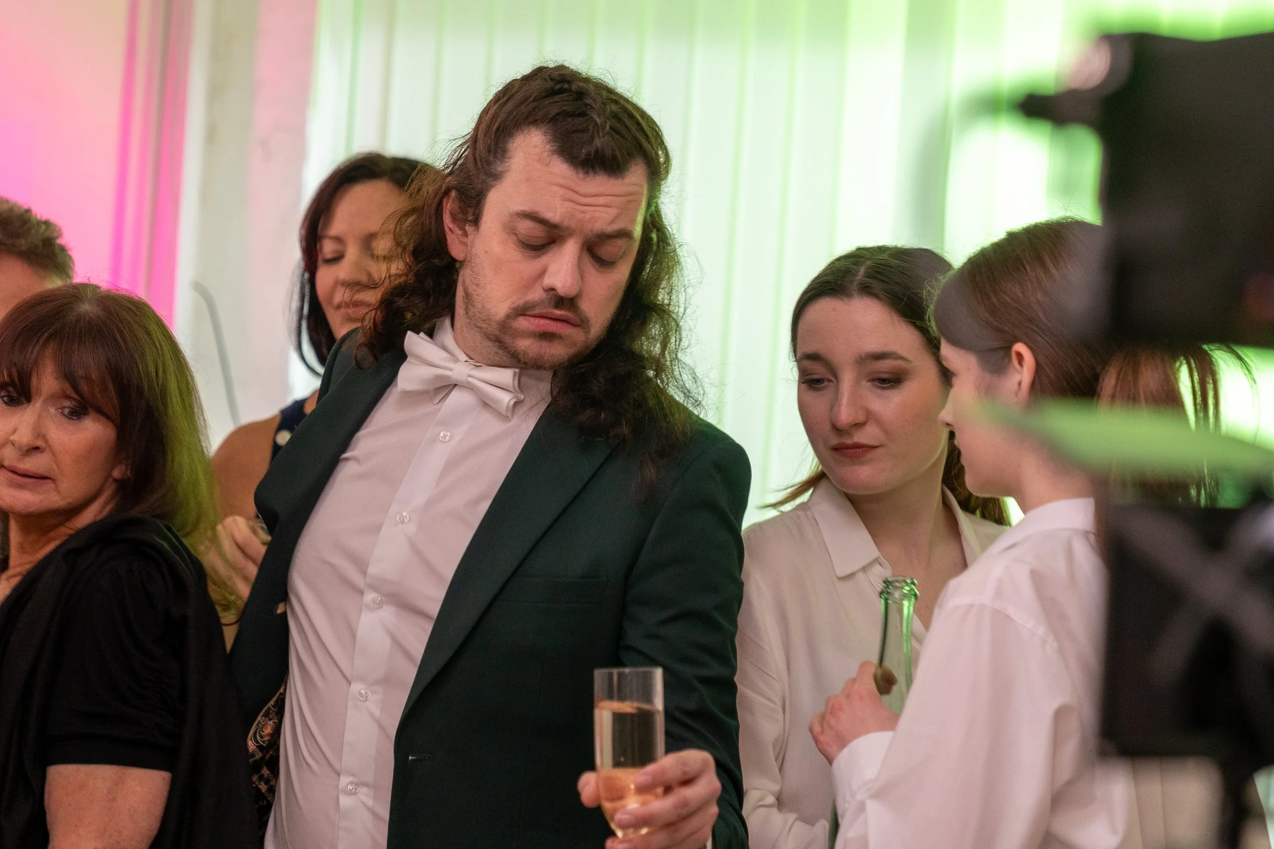 A man in a tuxedo holding a glass of champagne at a social gathering, surrounded by women dressed in formal attire, in a brightly colored room with pink and green lighting.