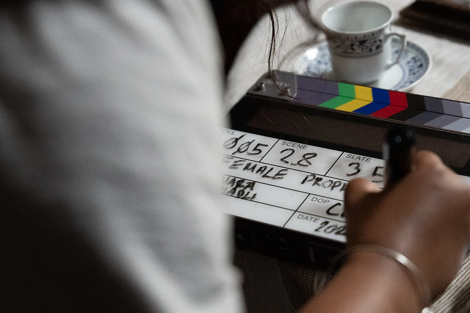 Close-up of a clapperboard with handwritten scene numbers, under a person's hand and arm, next to a tea cup and saucer on a wooden table.