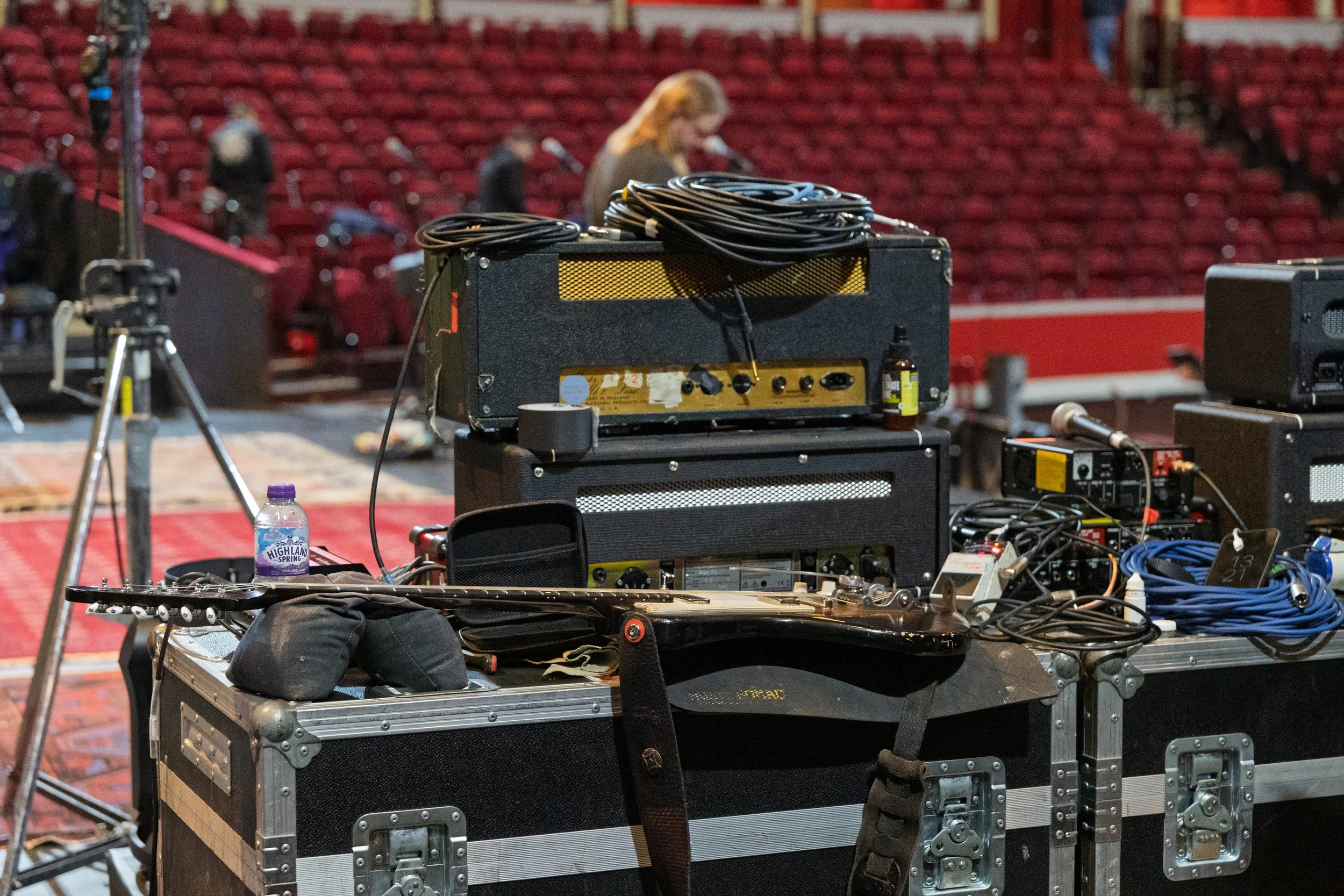 Musical equipment on stage, including guitar, amplifiers, cables, and a microphone, in an empty auditorium at the Royal Albert Hall with red seats.
