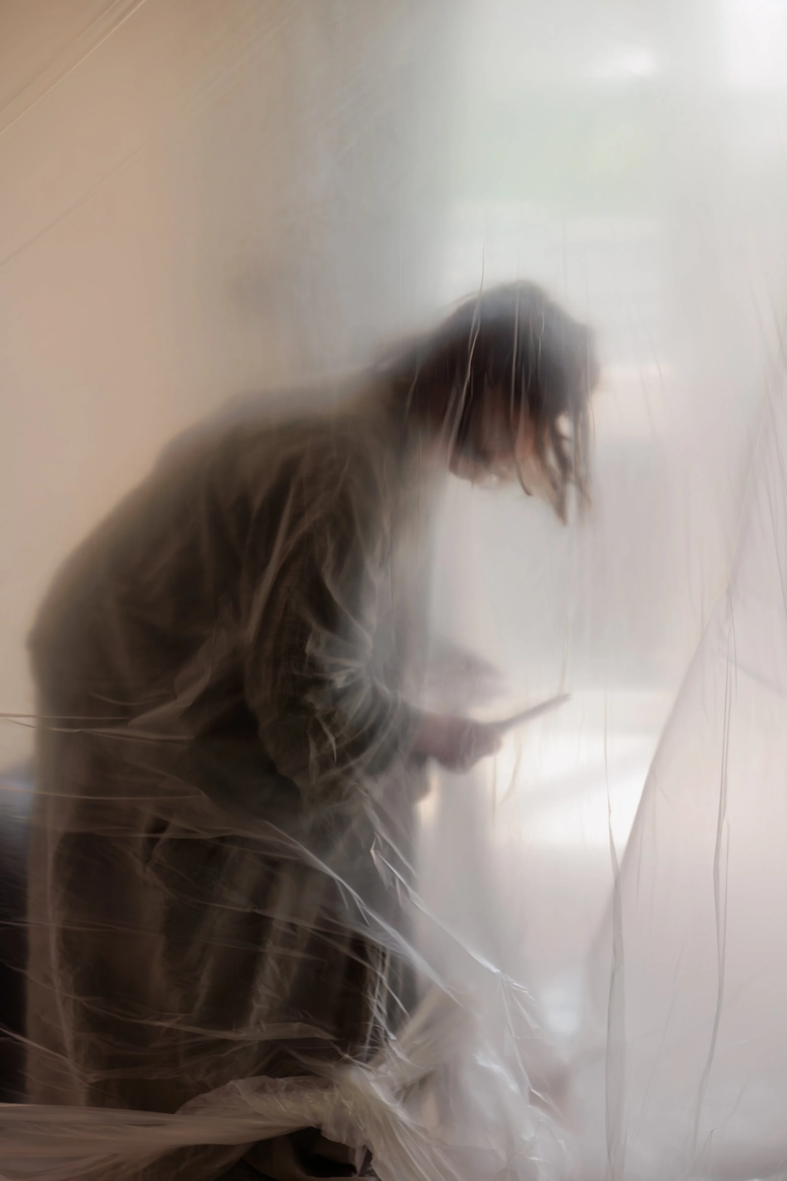 A person with dark hair and glasses is sitting behind a clear plastic curtain, with their head slightly down, in a room with plain light-colored walls.