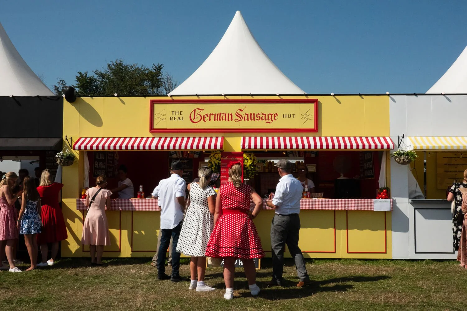 People standing in front of a yellow food stand with a red sign that reads 'German Sausage.' The stand has red and white striped awnings and is positioned in front of white tents on a clear day.