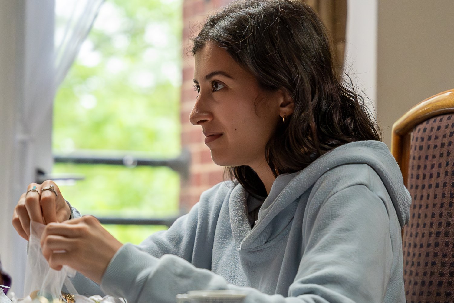 Young woman with dark hair and a light grey hoodie sitting at a table, opening a plastic bag, with a window showing green trees and a brick wall in the background. Film still.