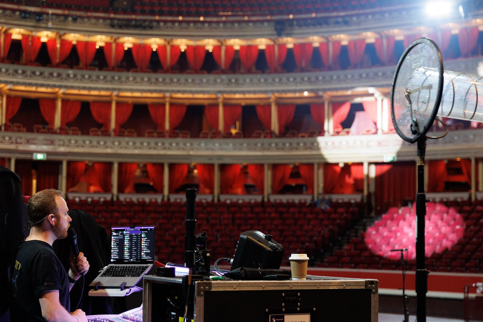 A man holding a microphone and working with a laptop at a sound control station inside a large theater with red curtains and multiple balconies.
