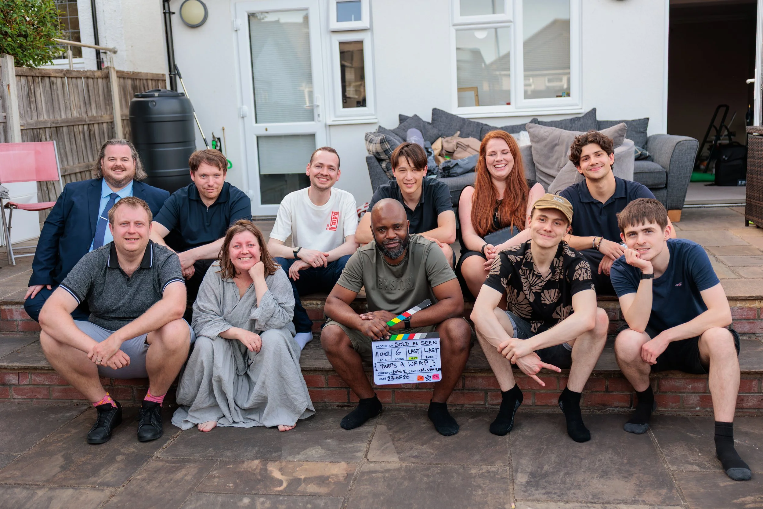 A group of eleven people sitting and kneeling on stone steps outside a house, smiling at the camera. There are six men and five women, with diverse appearances and expressions. One person in the front holds a clapperboard for 'Sold As Seen' (2026)