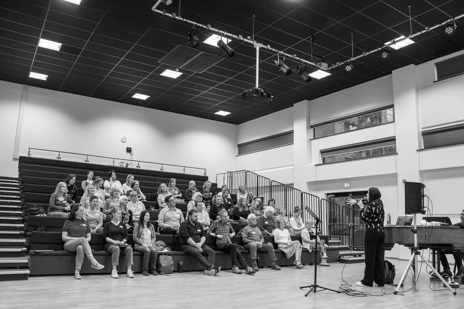 Black and white photo of a conductor addressing a choir in an auditorium.