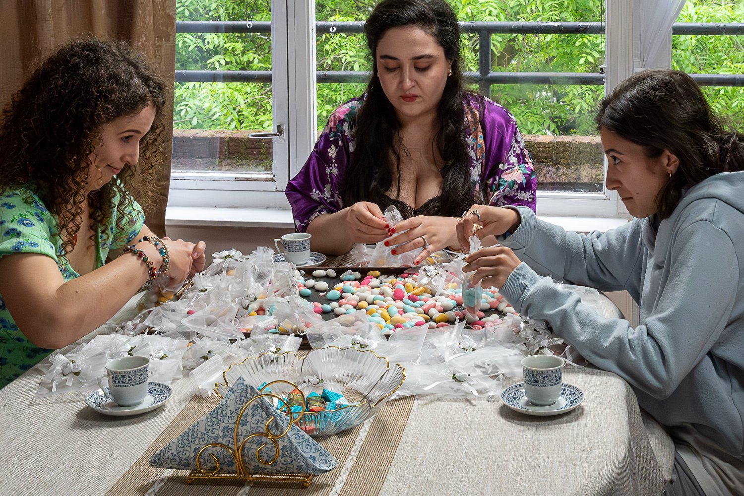 Three women sitting at a table with colourful heaps of sugared almonds, sorting them for a wedding, in a room with a large window showing green foliage outside. Film still.