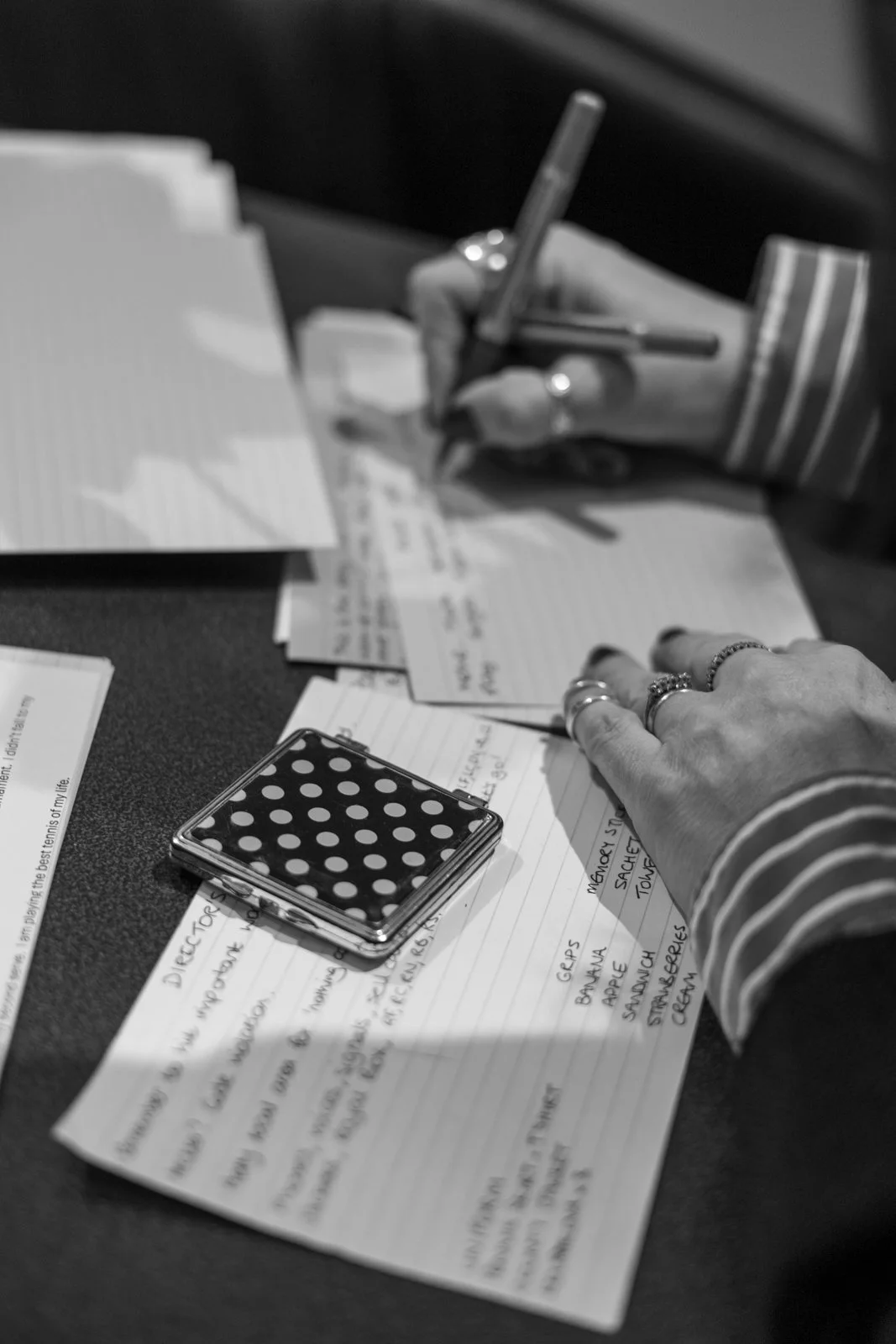 A person writing on lined cards with various notes and lists, with a polka-dotted compact mirror and several handwritten notes and papers on a dark surface. Comedian Pauline Eyre works on her script before the first date of her 'Anyone For Tennis?' t