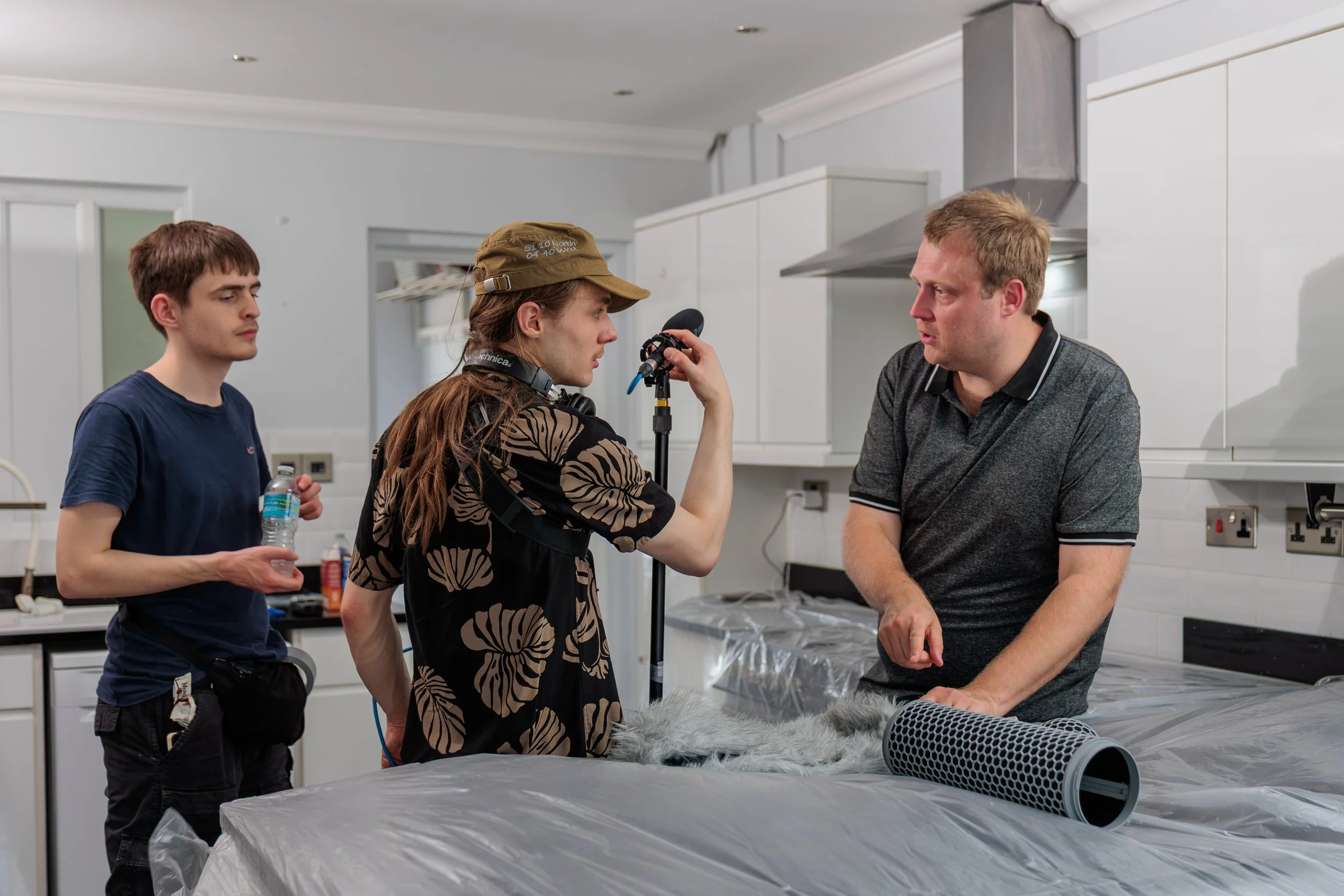 Group of four people in a room, two men and two women, engaged in a conversation or task, with a table covered in a plastic sheet and some equipment, possibly preparing for filming or a project.