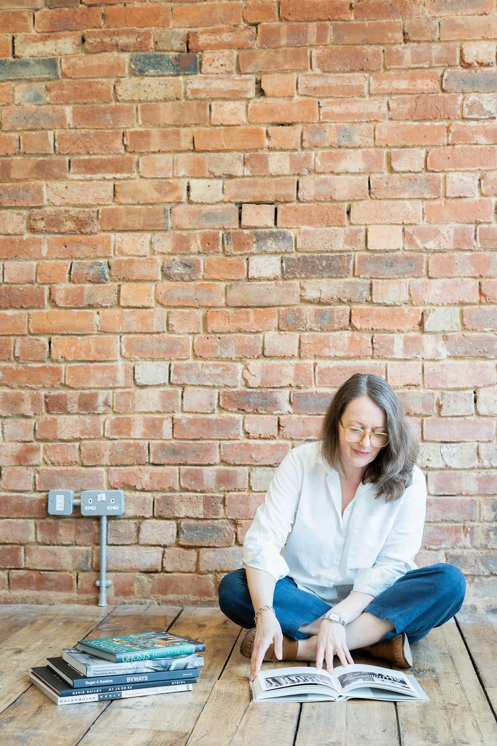 A woman with glasses and a white shirt sitting on a wooden floor reading a photo album, with a stack of books beside her, in front of a brick wall.