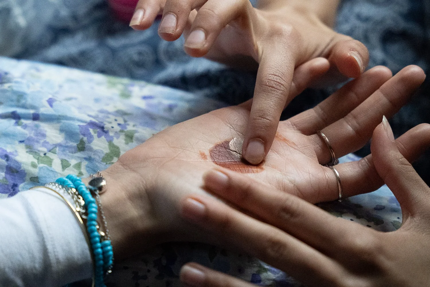 Close-up of two hands touching on a floral fabric surface, one hand with multiple jewelry pieces including turquoise beads, and the other hand with a metallic ring. The hand with jewelry has a tattoo of a leaf on the palm.