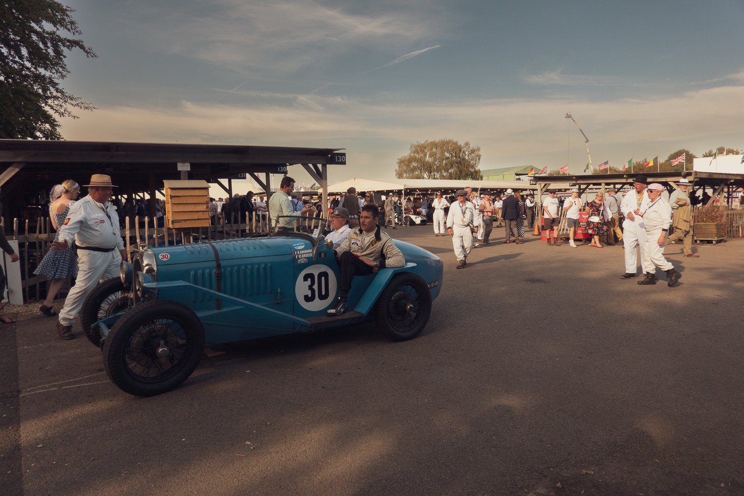 Vintage blue race car with the number 30, surrounded by people at a racing event, with tents and flags in the background on a sunny day.