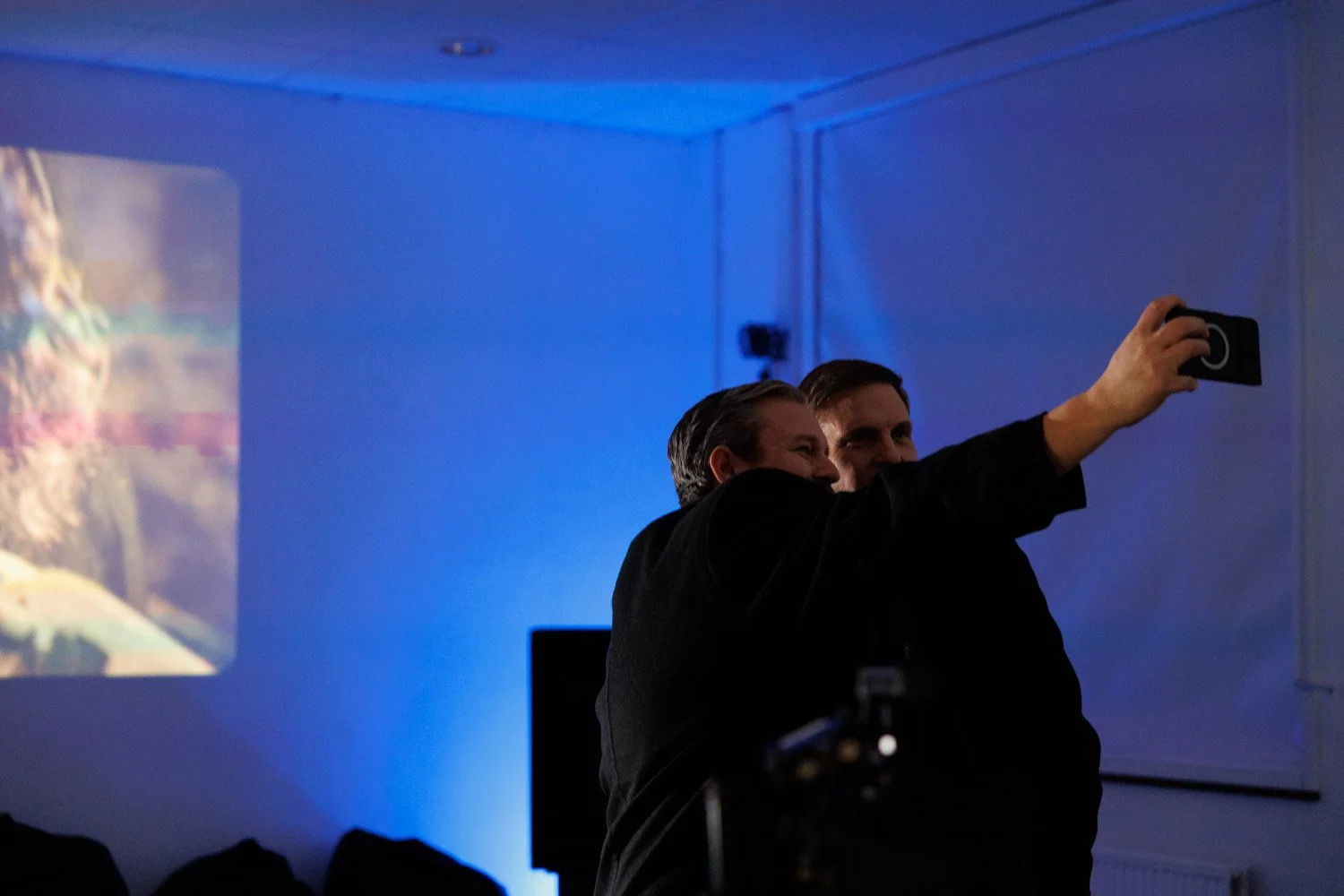 Two men taking a selfie in a dimly lit room with blue lighting and a projection on the wall