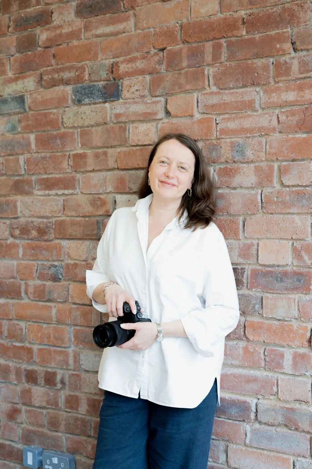 A woman with shoulder-length brown hair wearing a white button-up shirt and jeans, holding a camera in her right hand, standing against an exposed brick wall. It's Catherine, the photographer whose website you're reading. I’m