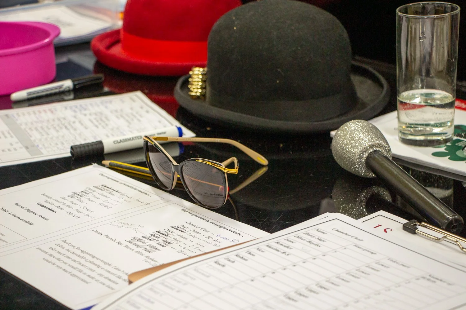 A table with a microphone, sunglasses, paperwork, a glass of water, a black hat on a hat stand, and miscellaneous items.