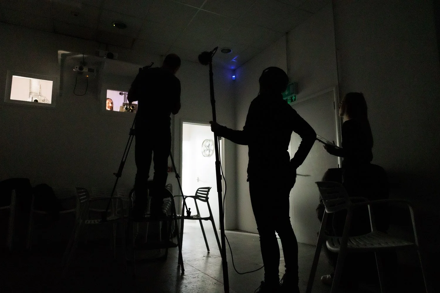 Silhouettes of three people in a dark room, possibly a filming or photography studio, with some chairs and a door visible, and two windows showing parts of a room with a white background.
