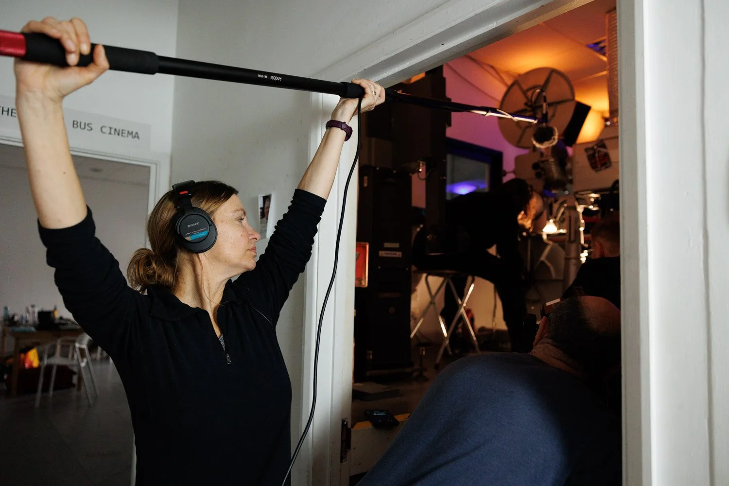 A woman with headphones holding a boom microphone over a scene being filmed inside a room with other crew members and equipment.