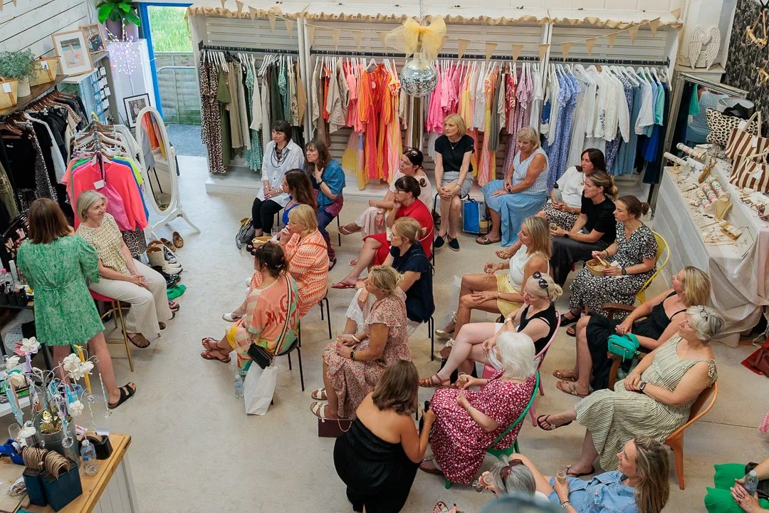 A group of women attending a make-up demonstration in a boutique, seated on chairs facing a woman speaker near a rack of colorful clothing.