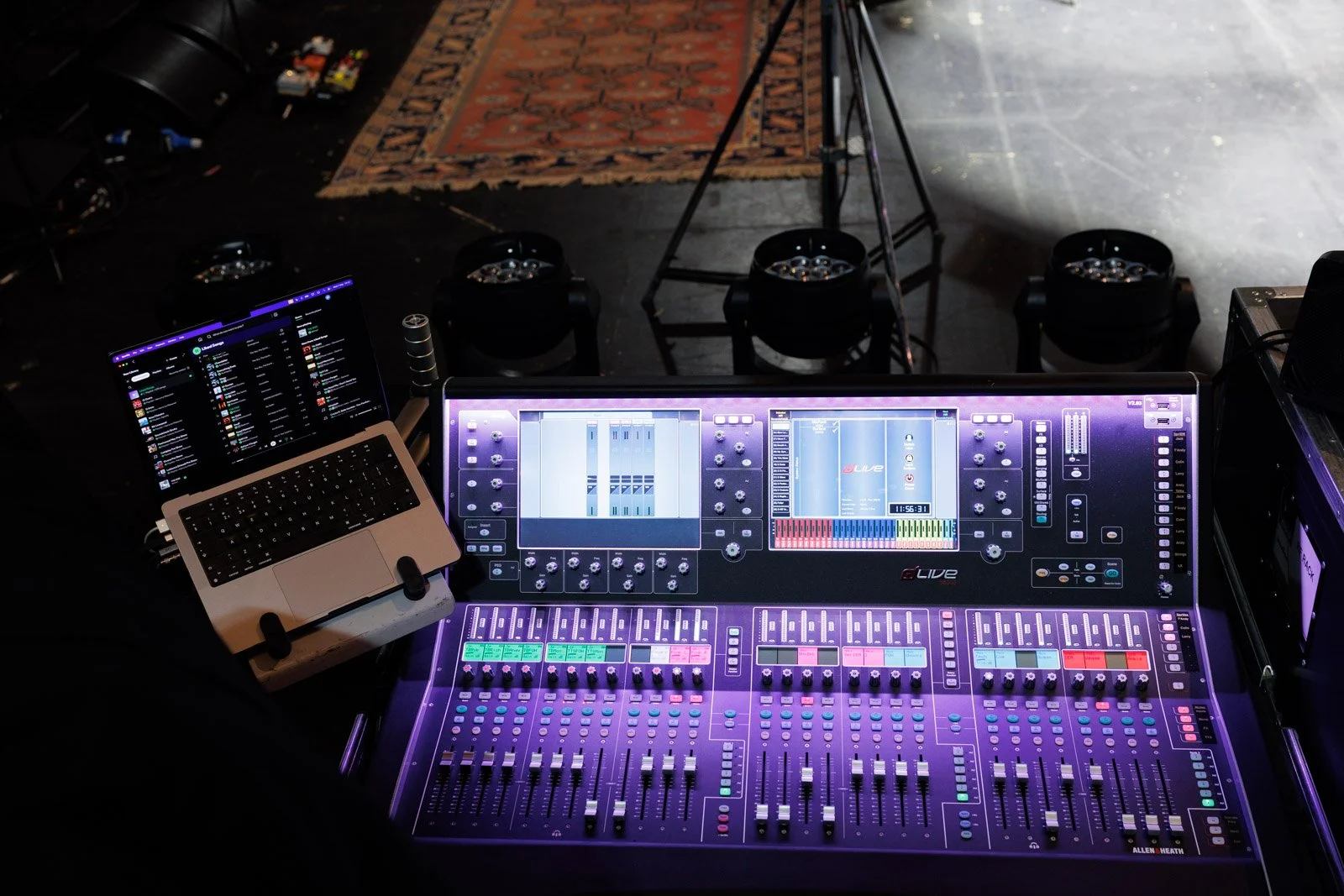 A professional audio mixing console with illuminated colorful controls, a laptop displaying a digital audio workspace, and a music playlist on a screen in a dark environment, likely a stage or recording studio.