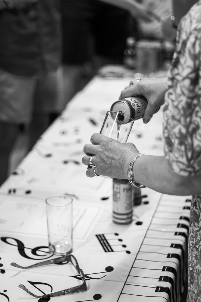 A woman is pouring a Pimms from a can into a glass on a table decorated with musical notes and a staff. She is wearing rings and a bracelet, and the table has glassware and a pair of glasses on it.