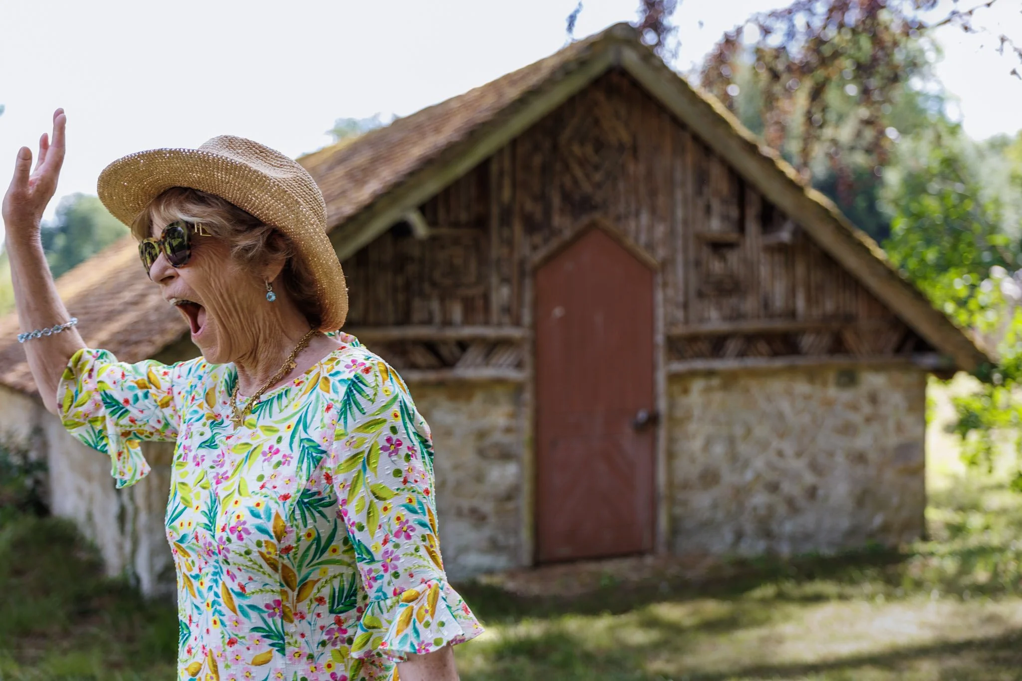 An elderly woman wearing a floral dress, sunglasses, and a straw hat, smiling and waving outdoors in front of a rustic wooden building surrounded by trees.