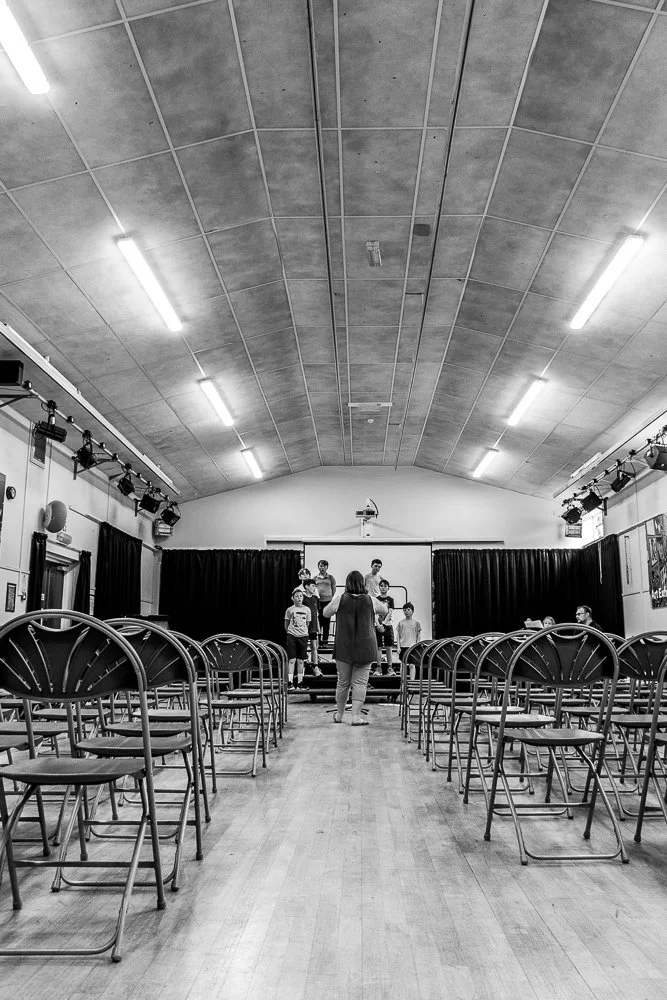 People rehearsing on stage in a small auditorium with rows of empty chairs, black curtains, and a projection screen.