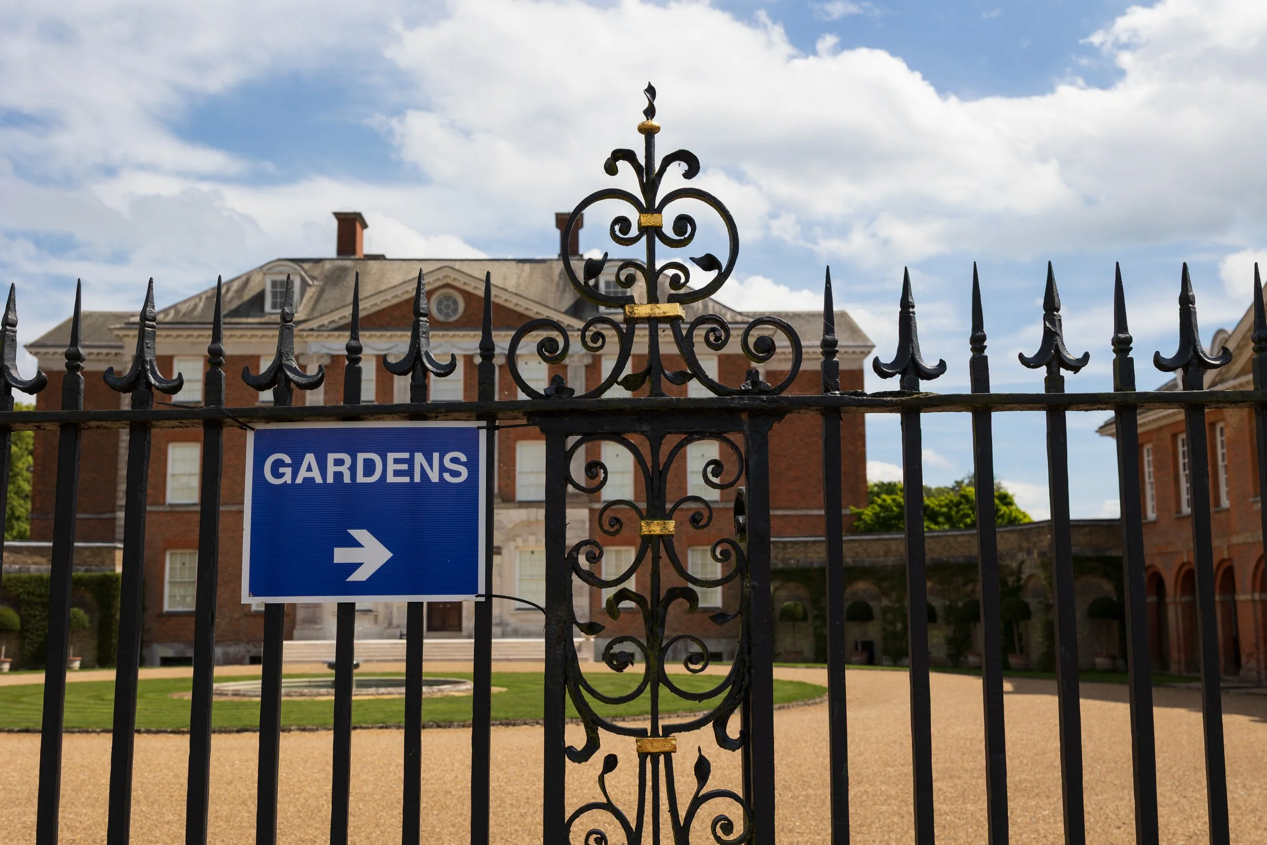 Ornate black metal gate with a travel sign pointing right towards gardens, in front of a red brick mansion with white accents and multiple windows, under a partly cloudy sky.