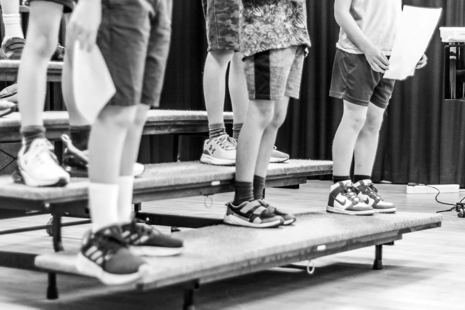 Children standing on risers, holding papers, practising a song before a concert.