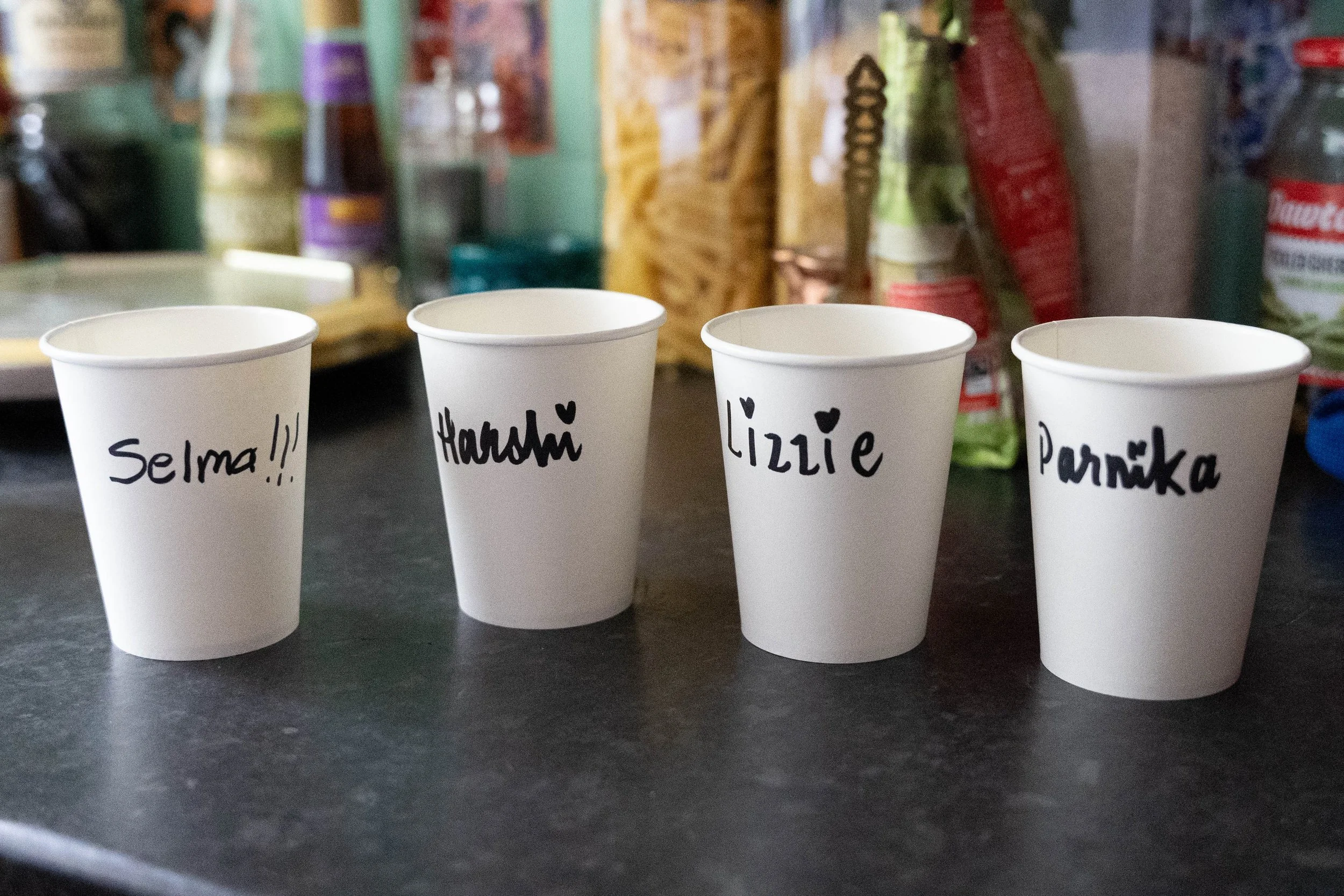 Four white paper cups with handwritten names in black marker, placed on a dark countertop. The cups are labeled 'Selma!!!', 'Harold♥', 'Lizzie', and 'Parrinka'. Background includes various food and drink items on shelves.