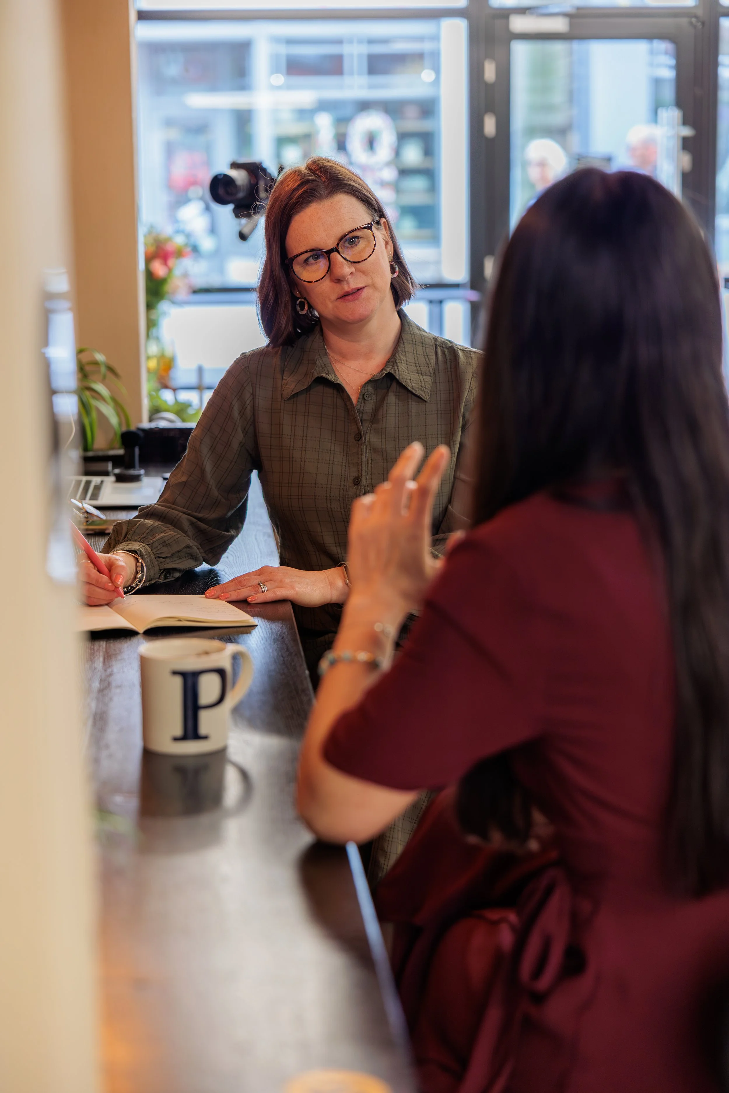 A woman with brown hair and glasses sitting at a counter in conversation with another woman whose back is to the camera. The woman with glasses has an open notebook and a pen in hand. There is a coffee mug with a letter 'P' on it on the counter, and 