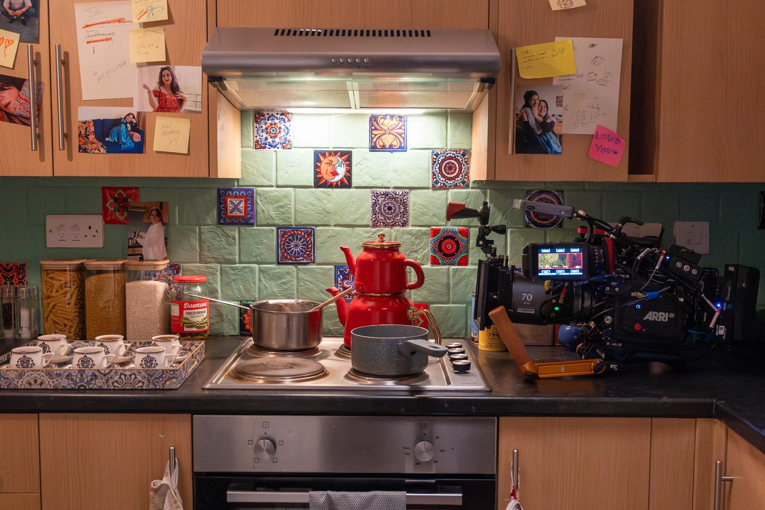 A kitchen stove with a red teapot, a small pot, and a saucepan on the burners. The backsplash has colorful decorative tiles, and the wall above is decorated with photos and notes on the cabinets.