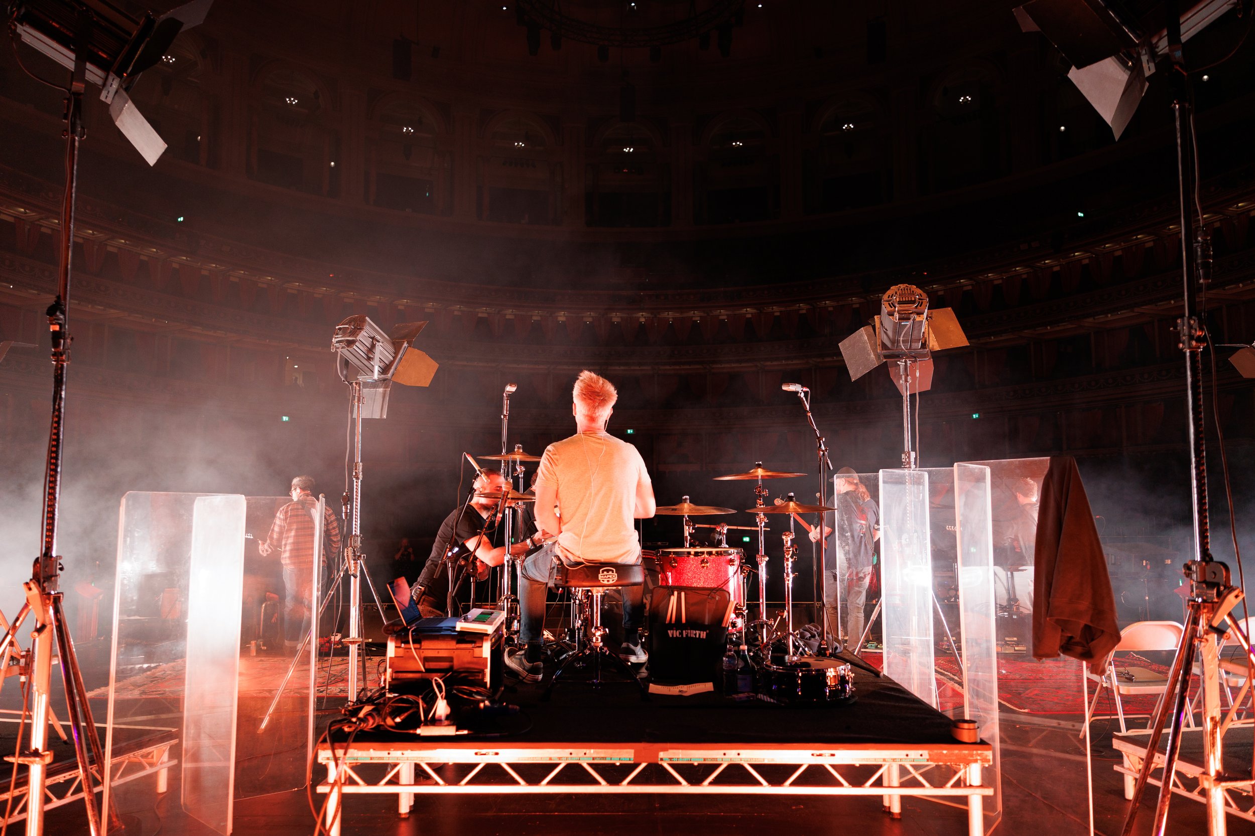 View from behind the stage showing musicians preparing for a performance in a large theater with ornate balconies. There are stage lights and equipment around them.