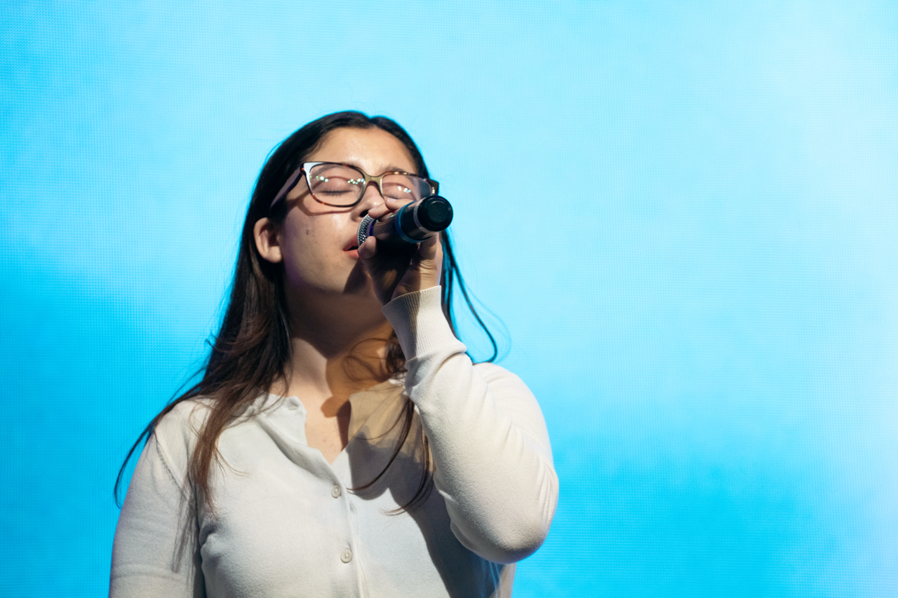 Person singing into a microphone with eyes closed against a blue background.