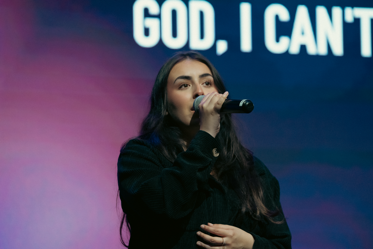 A woman singing into a microphone on stage with text "God, I Can'" behind her.