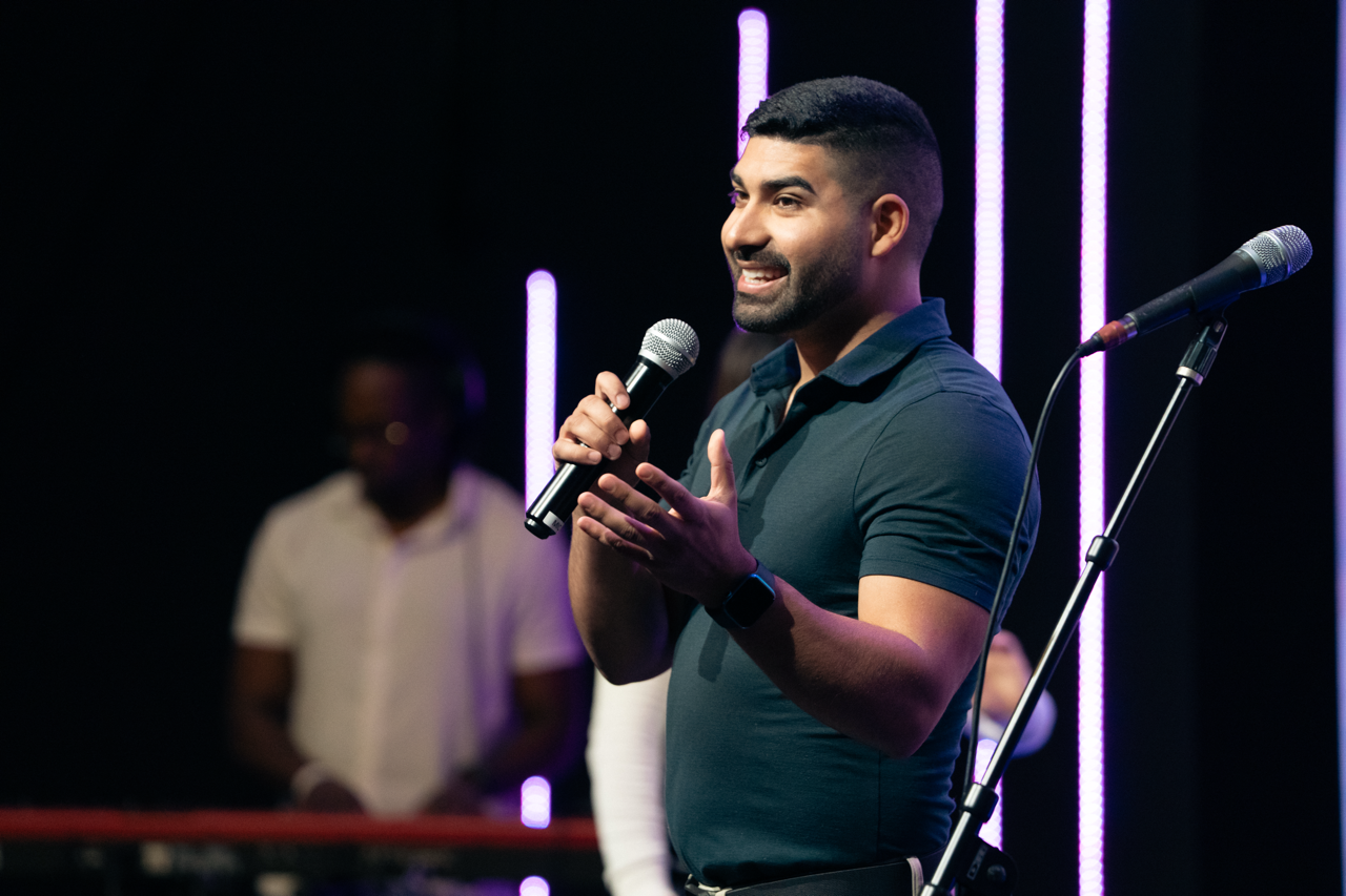 Man speaking into microphone on stage with purple lights and band in the background.