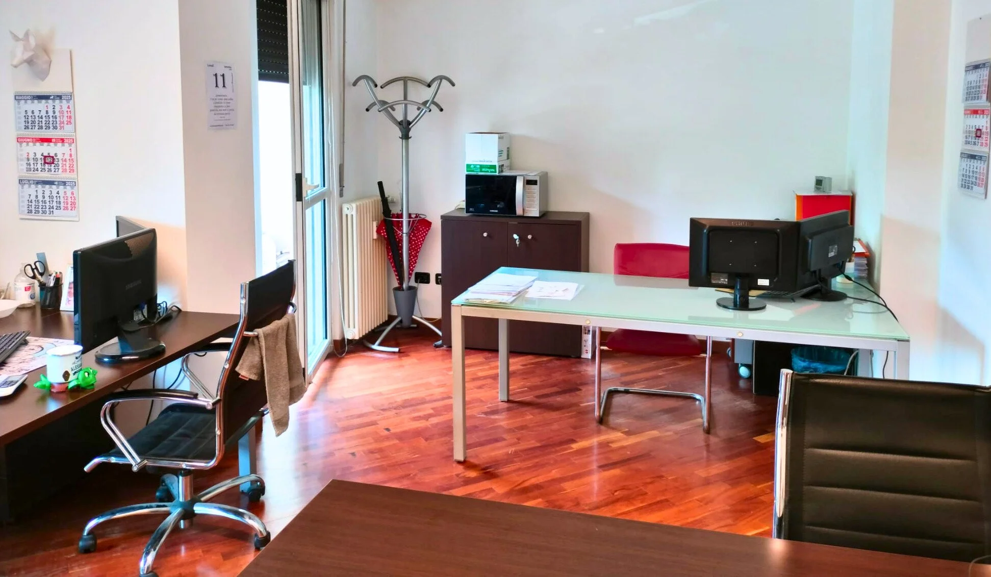 Ufficio con scrivanie, computer e sedie, calendari alle pareti, scaffale con documenti, e accessori come ombrelli e piantine, pavimento in legno.