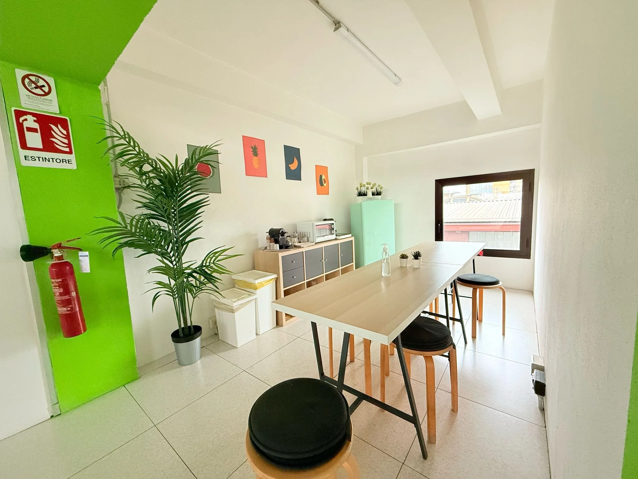 Interno di una stanza con pareti bianche e arredi moderni. Tavolo in legno con sedie e alcuni vasetti di piante, finestra grande con vista sulla città, decorazioni con quadri di frutta, cucina con microonde e dispensa. Parete verde con estintore e segnale di divieto di fumo.