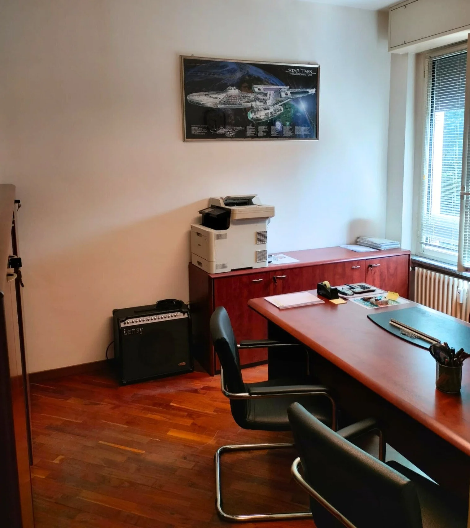 Ufficio con scrivania, sedie nere, stampante, lampada aerea, poster di Star Trek sulla parete e una finestra con tapparelle.