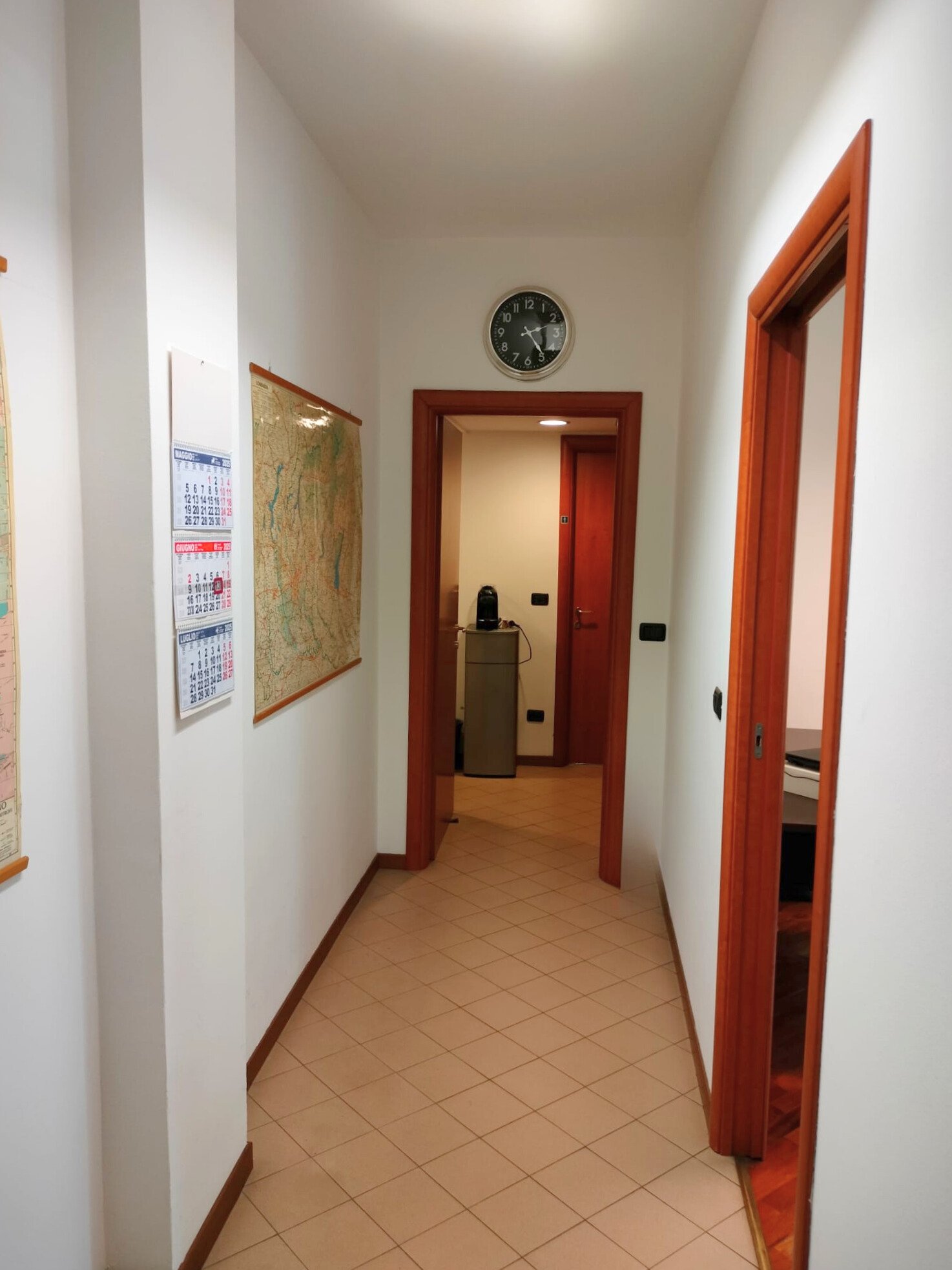 Corridor con pareti bianche, porte in legno, orologio da parete nero e bianco, mappe e calendari sui muri, pavimento in piastrelle beige