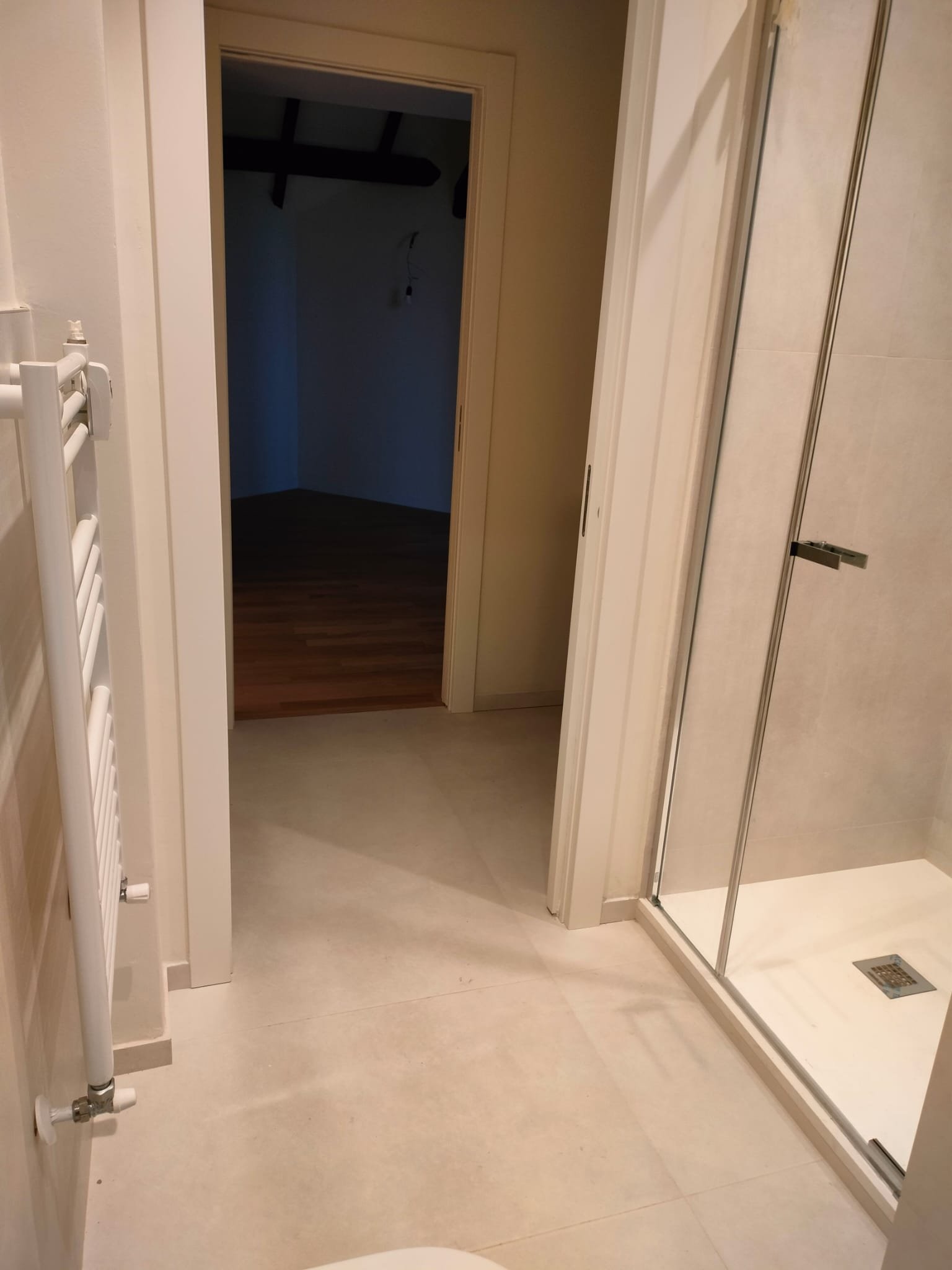 In bagno, vista della porta aperta verso una stanza buia, con un piatto doccia in vetro e piastrelle beige.