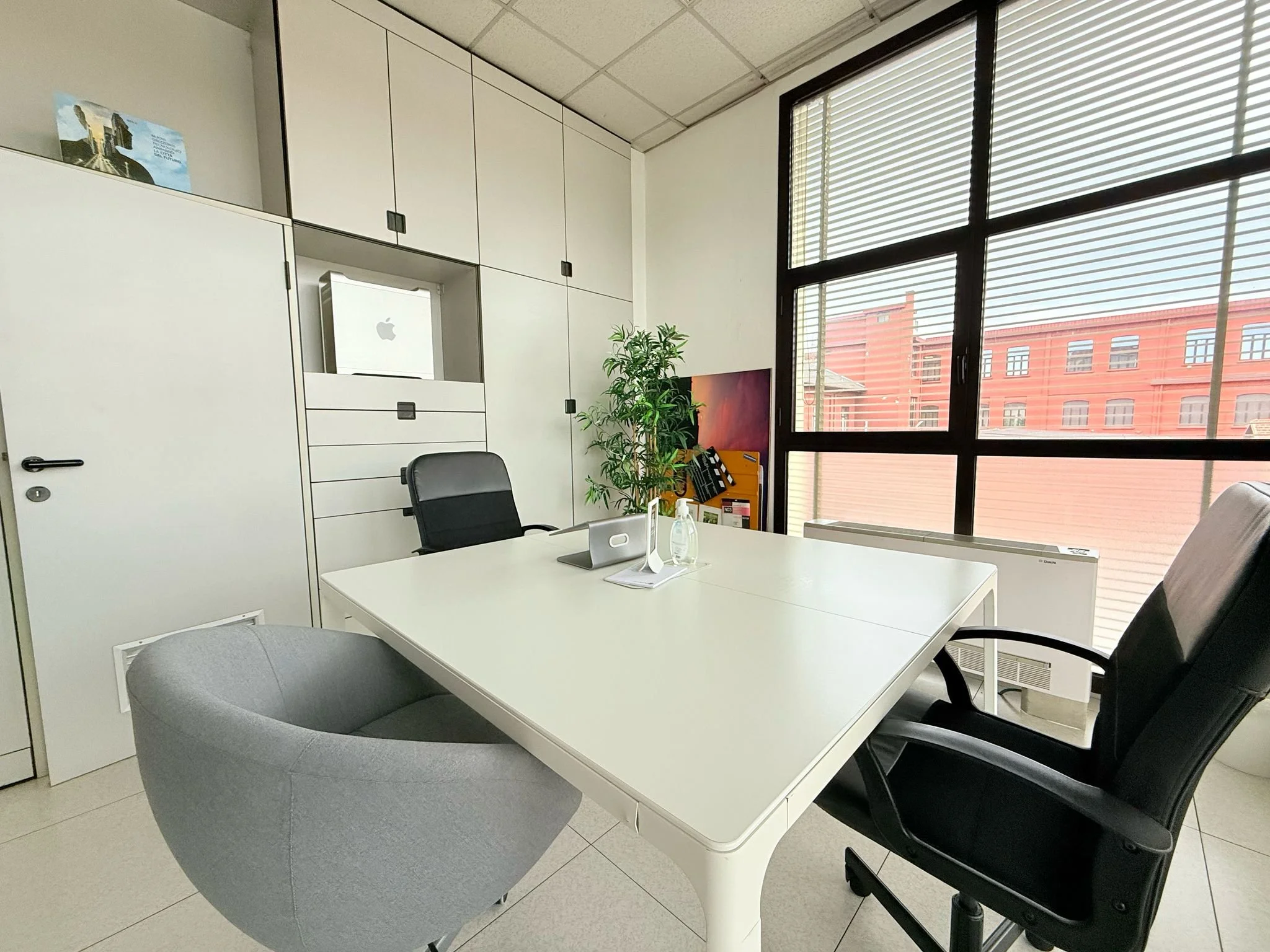 Ufficio luminoso con grande finestra, scrivania bianca con tre sedie (una nera, una grigia, una gialla), piante verdi e armadietti bianchi.