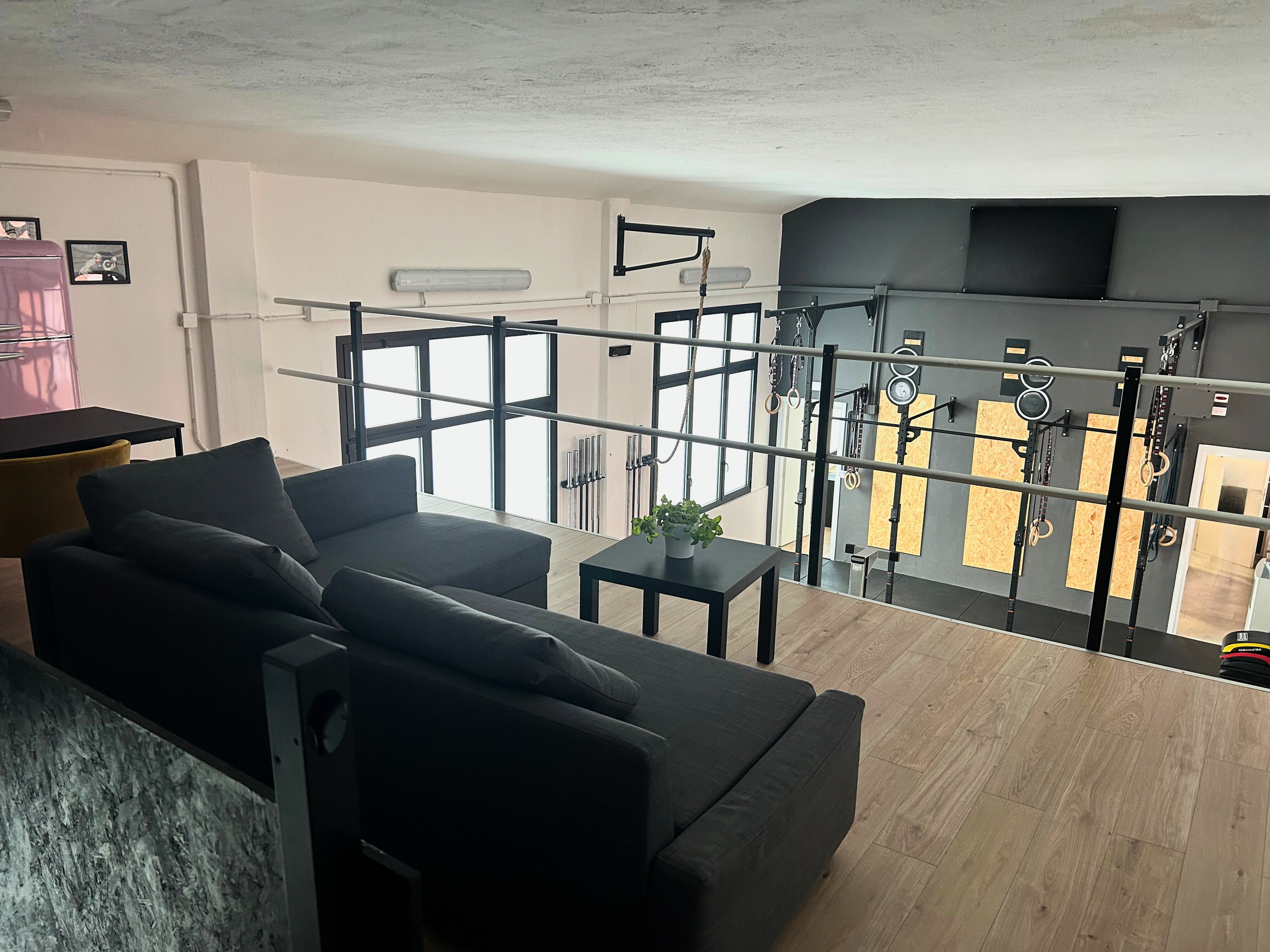 Zona living di uno spazio fitness con divano, tavolino e palestra sullo sfondo, luce naturale dalle finestre.