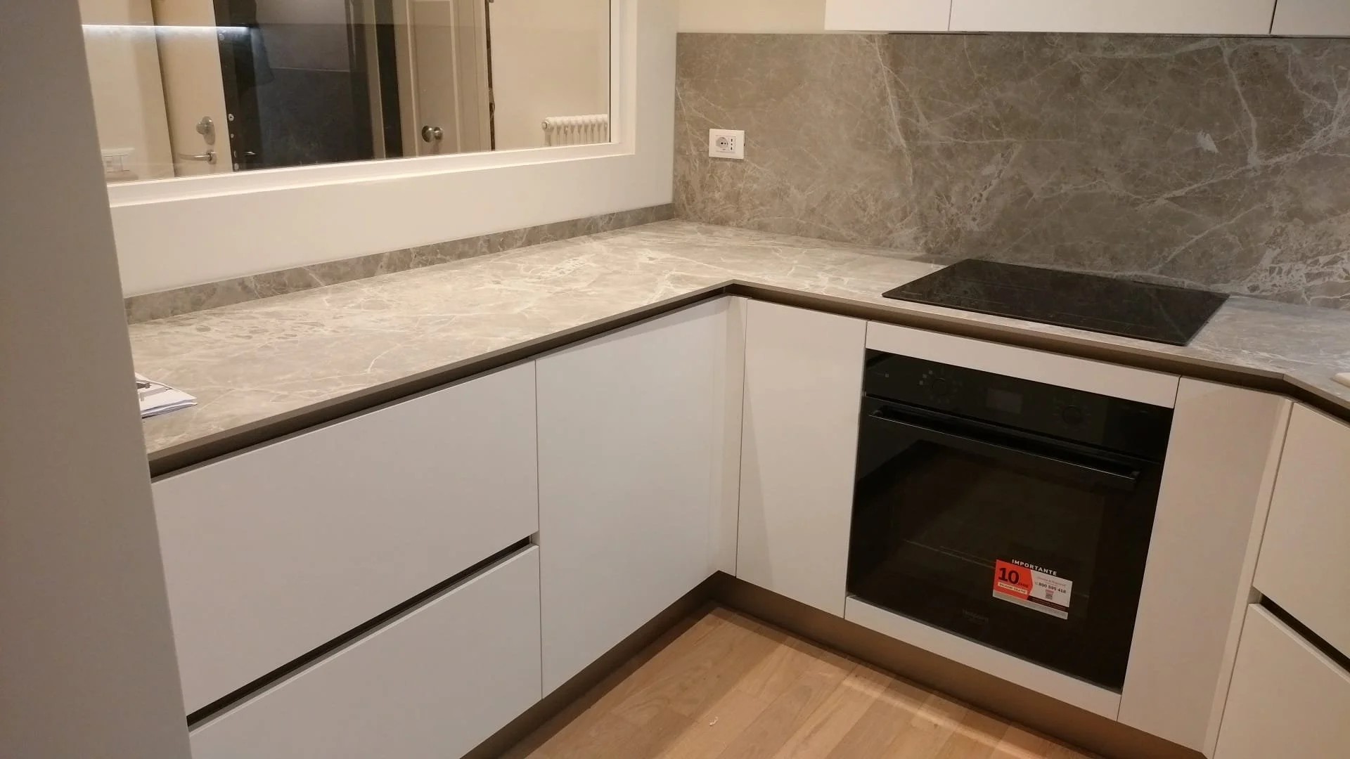 Angolo di cucina con mobili bianchi, piano in marmo beige, forno nero e piano cottura in vetro nero. C'è una spina elettrica sulla parete e un finestrino sopra il piano di lavoro.